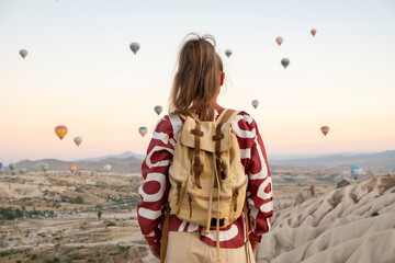 Hot air balloons float in the sky as a traveler stands on a hill in Cappadocia during sunrise observing the view in Turkey