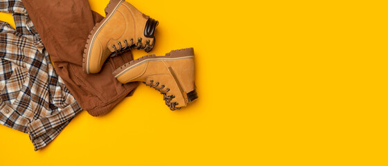 Fashionable clothes for child, boy, teenager. Yellow boots made of genuine leather nubuck, plaid shirt, brown trousers on yellow background. Rough autumn-winter work boots with ribbed sole and laces 