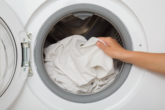 Young adult woman hands holding and unloading white sheet from opened washing machine. Clothes care. Closeup. Front view.