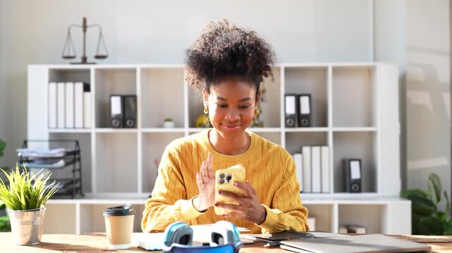Smiling young african american female lawyer sitting at her desk and texting with her smartphone while taking a break from work in a modern bright law firm office with a scales of justice