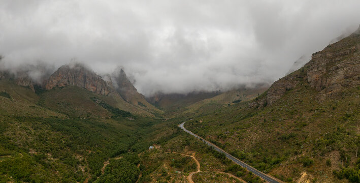 Aerial view of a winding road snakes through the verdant valley, cradled by rugged mountains veiled in misty clouds, creating a serene yet dramatic landscape, Paarl, Western Cape, South Africa.