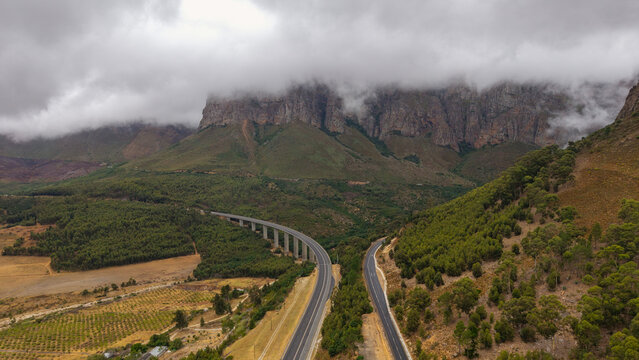 Aerial view of a modern viaduct and road cutting through the verdant valley beneath cloud-topped mountains, a symphony of grey and green, Paarl, Western Cape, South Africa.