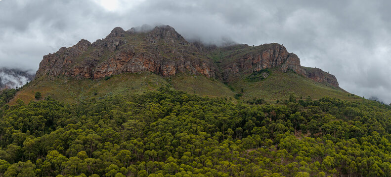 Aerial view of the rugged mountain rises majestically, its rocky face a tapestry of browns and oranges, crowned by swirling mist, Paarl, Western Cape, South Africa.