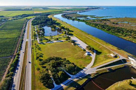 Aerial view of the vibrant green park area beside a long highway and winding waterway, contrasting with the surrounding landscape, Lake Okeechobee, John Stretch Memorial Park, Florida, United States.