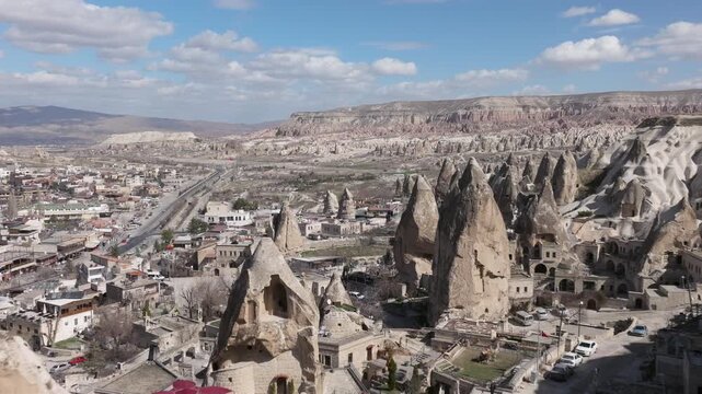 Aerial drone footage of G&ouml;reme town in Cappadocia, Turkey, surrounded by fairy chimneys and volcanic rock formations. The shot reveals traditional stone houses, cave dwellings carved into soft tuff ro