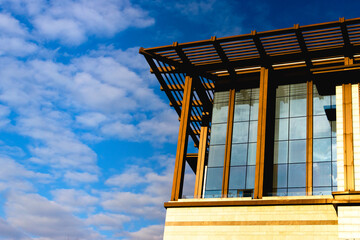 Modern architectural structure with overhanging slatted roof, vertical beams, large glass windows and light stone base, vivid blue sky with scattered clouds and contemporary facade detail.