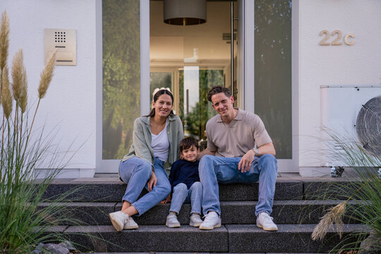Family sitting together in front of modern home with heat pump system