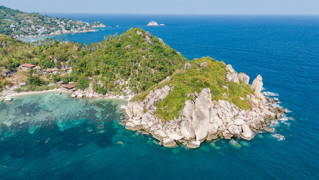 Aerial view of a lush, green island with rocky outcrops meeting the turquoise sea, contrasting with the distant shoreline, Ko Tao, Surat Thani, Thailand.