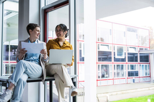 Coworkers discussing work by office window with laptop and documents