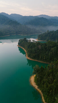 Aerial view of the turquoise waters of Cheow Lan Lake embrace the lush, emerald green rainforest, a serene vista of nature's grandeur, Khao Sok National Park, Surat Thani, Thailand.