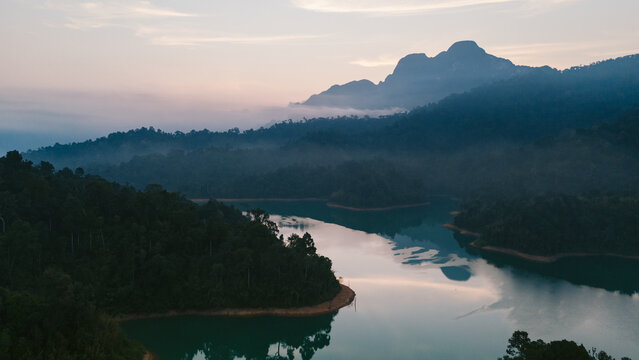 Aerial view of a serene lake mirroring the misty mountains and verdant forest under a pastel sky, Khao Sok National Park, Surat Thani, Thailand.