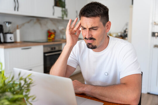 Concentrating man working from home on laptop experiencing severe headache, feeling stressed and frustrated with work, suffering from migraine and pain