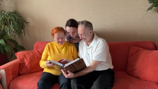 Joyful attractive senior woman and cheerful handsome mature man sitting on couch, watching family photo album, sharing and remembering happy and funny moments while retired couple relaxing.