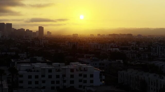 High angle aerial view of Los Angeles at golden hour with sun rays visible through haze, downtown skyline left, CVS and illuminated signs visible, white residential buildings in shadow, palm trees