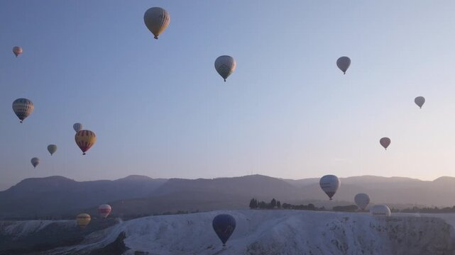 Aerial drone footage of colorful hot air balloons flying above the white travertine terraces of Pamukkale, Turkey, at sunrise. The balloons float over the mineral formations and thermal pools, creatin