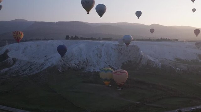 Aerial drone footage of colorful hot air balloons flying above the white travertine terraces of Pamukkale, Turkey, at sunrise. The balloons float over the mineral formations and thermal pools, creatin