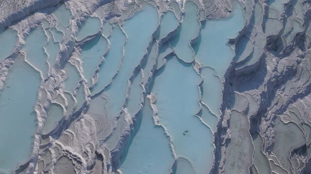 Aerial drone footage of the white travertine terraces of Pamukkale in Turkey, filled with natural thermal water pools. The bright mineral formations create a striking contrast with the surrounding lan