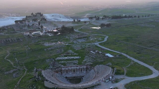 Aerial drone footage of the ancient Roman amphitheater of Hierapolis overlooking Pamukkale, Turkey, at sunset. The well-preserved stone structure stands on a hillside above the famous white travertine