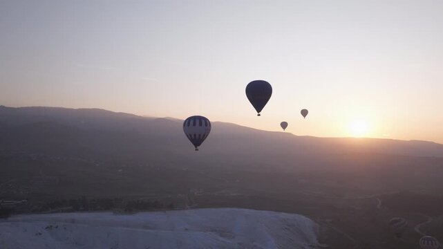 Aerial drone footage of colorful hot air balloons flying above the white travertine terraces of Pamukkale, Turkey, at sunrise. The balloons float over the mineral formations and thermal pools, creatin