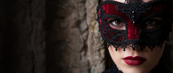 Mysterious woman in elegant red velvet and black lace Venetian mask. Dramatic masquerade portrait on dark stone background for Carnival concept with copy space.