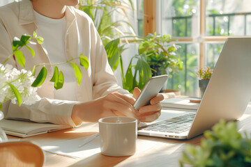 Person checks phone while working at home office with laptop and plants around in a bright room during daytime