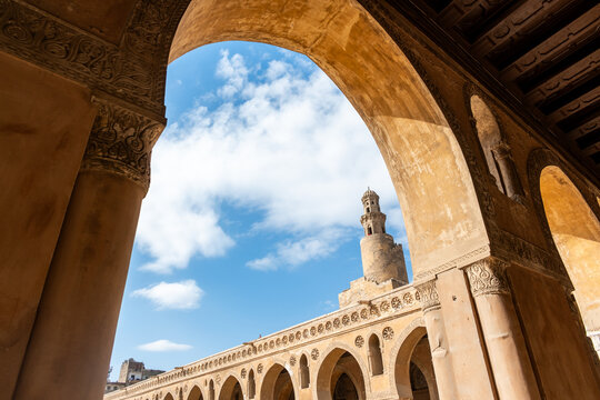 Ibn Tulun Mosque in Cairo viewed through arches. The mosque features a tall minaret and intricate architectural details. Blue sky visible in the background.