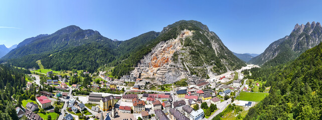 Aerial view of a mining town nestled beneath the stark, scarred mountainside contrasting with lush green forests, Cave del Predil, Friuli-Venezia Giulia, Italy.