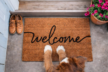 A dog stands on a welcome mat at the entrance of a home with a pair of shoes and a flower pot