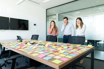Multicolored sticky notes covering conference table with three business people laughing in background. Creative brainstorming and teamwork concept for office.