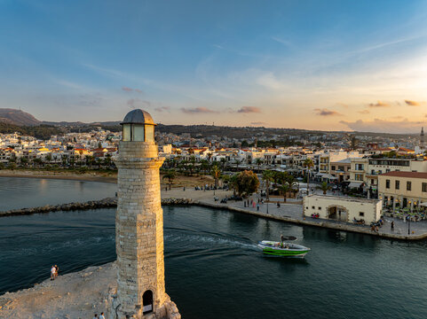 Aerial view of the old lighthouse standing guard as a vibrant green boat glides through the harbor waters, embraced by the historic architecture of Rethimno, Rethymno, Greece.