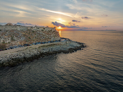 Aerial view of the sun setting behind the Fortezza of Rethymno as it casts a golden glow over the dark, rippling sea, Rethimno, Rethymno, Greece.