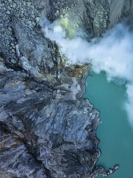 Aerial view of the turquoise lake meeting the textured, rocky landscape, with plumes of white smoke rising into the atmosphere, Banyuwangi, East Java, Indonesia.
