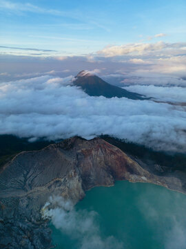 Aerial view of the Kawah Ijen volcano's turquoise crater lake steaming gently amidst the stark, rugged landscape contrasting with the soft, enveloping clouds, Banyuwangi, East Java, Indonesia.
