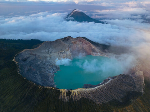 Aerial view of the turquoise lake nestled within the volcanic crater rim, framed by ethereal clouds and distant mountain peaks, Banyuwangi, East Java, Indonesia.