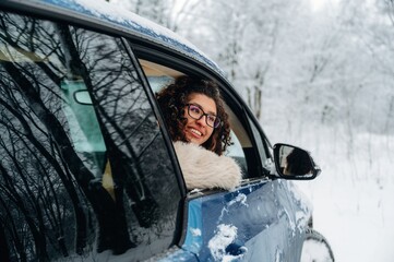 Young woman with curly hair wearing glasses is smiling from the window of a blue car covered in snow, surrounded by a winter forest landscape with trees in the background