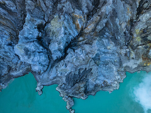 Aerial view of the rugged terrain meeting the turquoise waters, a stark contrast of textures and tones, Kawah Ijen, East Java, Indonesia.