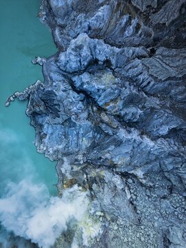 Aerial view of jagged, rocky cliffs meet a turquoise lake, while wisps of white smoke curl upwards, creating a mesmerizing contrast of textures and tones, Banyuwangi, East Java, Indonesia.