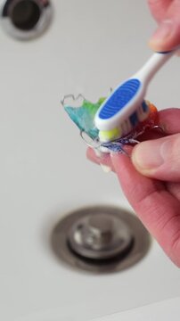 Hand cleans colorful orthodontic retainer with toothbrush and toothpaste in bathroom sink, closeup daily dental hygiene.