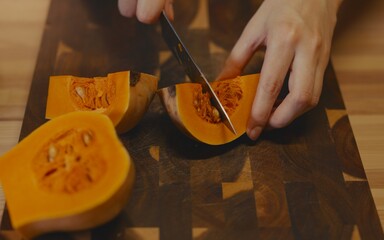 Female hands slicing halves of a butternut squash into smaller quarters on an acacia wood cutting board. Step-by-step healthy meal preparation, cooking process in a bright, rustic kitchen setting.