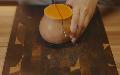 Close-up of female hands using a sharp knife to cut a ripe butternut squash in half on a wooden butcher block cutting board. Professional kitchen food preparation, organic healthy cooking concept.