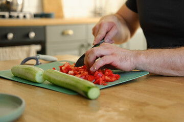 Man preparing fresh vegetables in modern kitchen, organic ingredients. Healthy meal. Cooking Food At Home. Male slicing red pepper and green cucumber on a wooden cutting board. Nutritionist, culinary