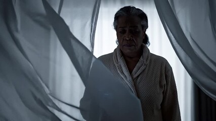 A dark-skinned woman stands in a dimly lit room with intimate video blackmail threateningly looming from behind a semi-transparent white curtain.