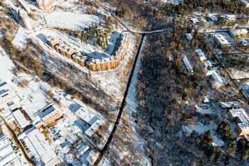 Winter aerial view of a vibrant urban parkland transition, featuring a modern curved building amidst pristine snow and bare trees, bathed in crisp sunlight
