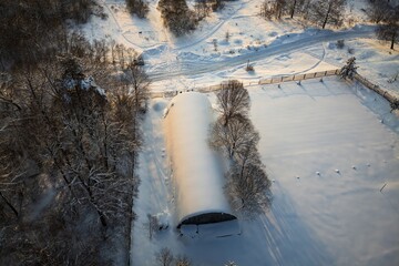 Aerial drone shot showcasing a pristine snow-covered hangar, its arched roof glowing under gentle winter sunlight. Surrounded by frosted trees and vast white terrain, a tranquil scene