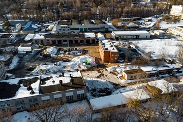 Aerial view of a snowy industrial zone, with a mix of old and new buildings, construction sites, and bare trees, under a clear winter sky