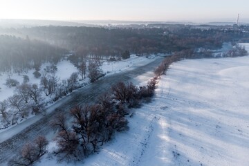 Aerial view of a serene winter river at dawn, framed by snow-covered banks and bare trees, with a hint of distant mist and a hazy horizon