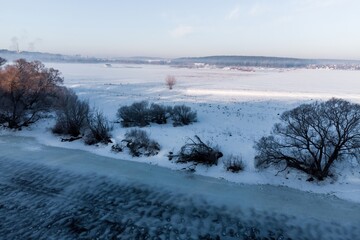 A tranquil winter morning unfolds along a frozen river, with bare trees silhouetted against a pale sky, their branches dusted with frost, creating a serene, frosty landscape