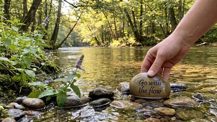 Hand places inscribed stone reading 'Go with the flow' into a sunlit river bend amidst a tranquil forest.