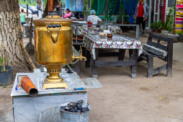 Brass samovar sits elegantly on a patterned tablecloth at an outdoor cafe. Blurred figures in background create a relaxed, inviting atmosphere during daytime.