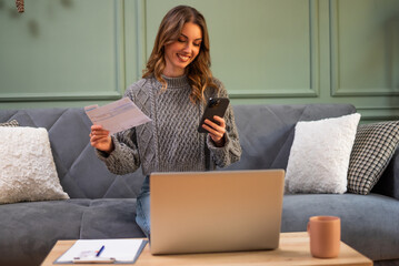 Happy adult woman is holding bills in her hand and using smartphone and laptop to pay online.